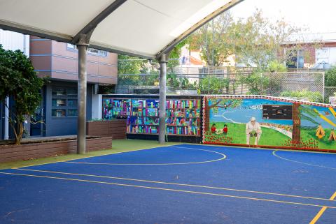 A blue turf basketball court with a mural at one end, covered with a roof but outside